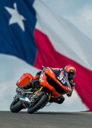 kyle wyman leans around a corner with giant texas state flag in background