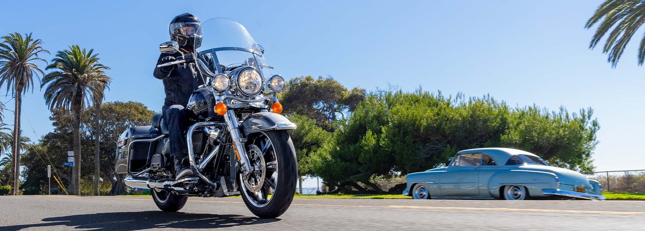 Motociclista con indosso l’equipaggiamento Harley in nero, alla guida di una Road King 2022 su per una strada di montagna, con la città sullo sfondo
