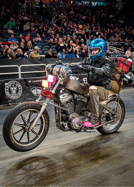 Chopper motorcycle packed for long distance camping does a wheelie in front of a cheering arena crowd