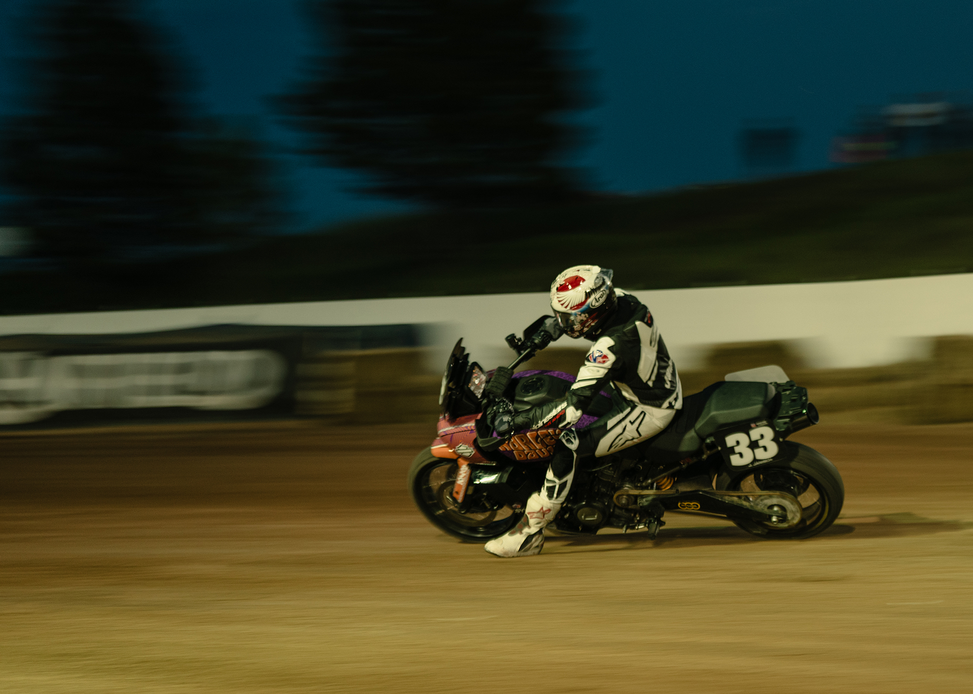 Motorcycle racer in full gear leans into a turn on a dirt track at night.