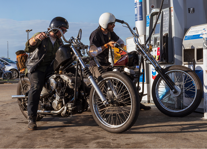 Two riders on custom motorcycles side by side getting gas at a gas station