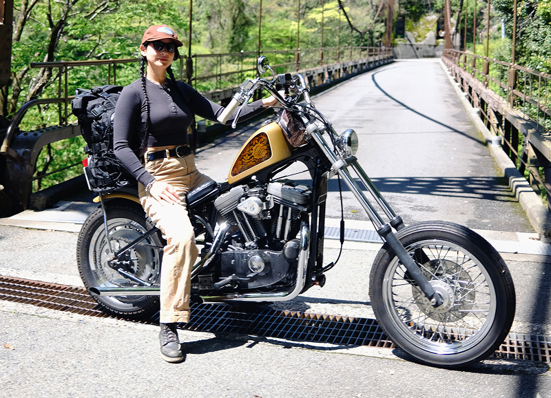 Adri Law sitting on a custom motorcycle parked in front of a small rural bridge with greenery along the path
