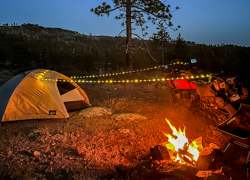 Night campsite with glowing tent, campfire, and Red H-D Pan America motorcycle under string lights in forest