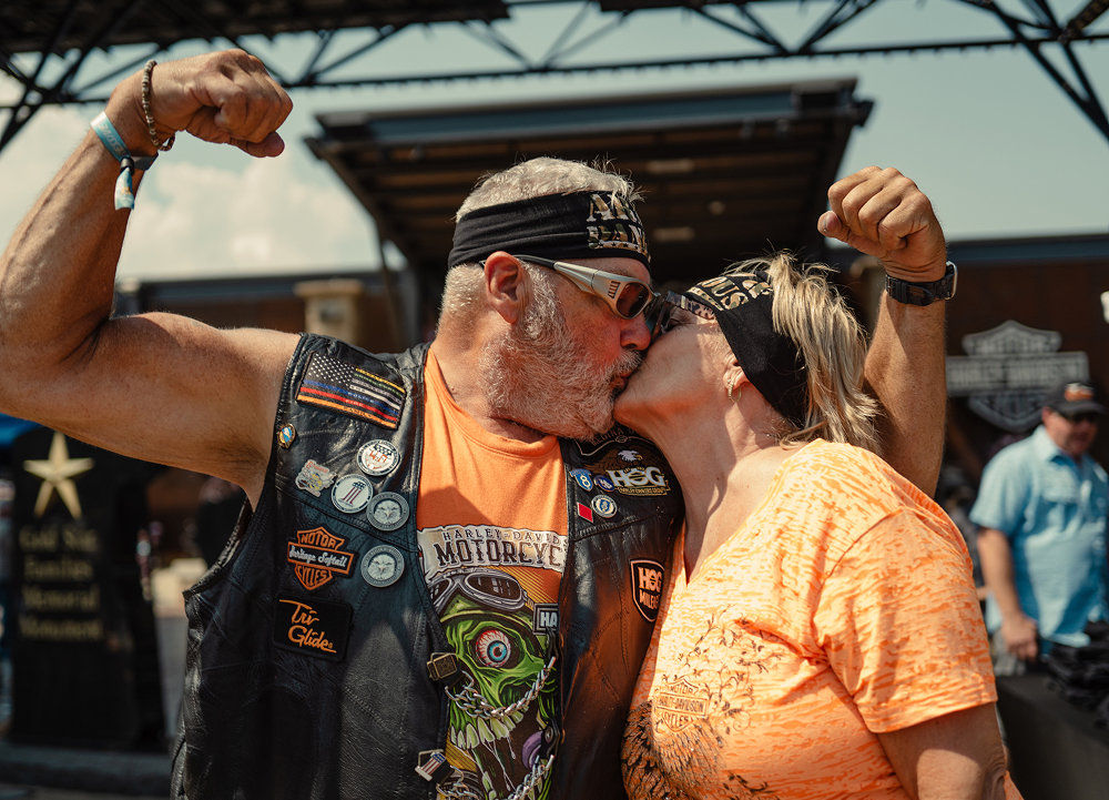 Two motorcycle rally attendees share a kiss while posing beside vendor displays and event crowds