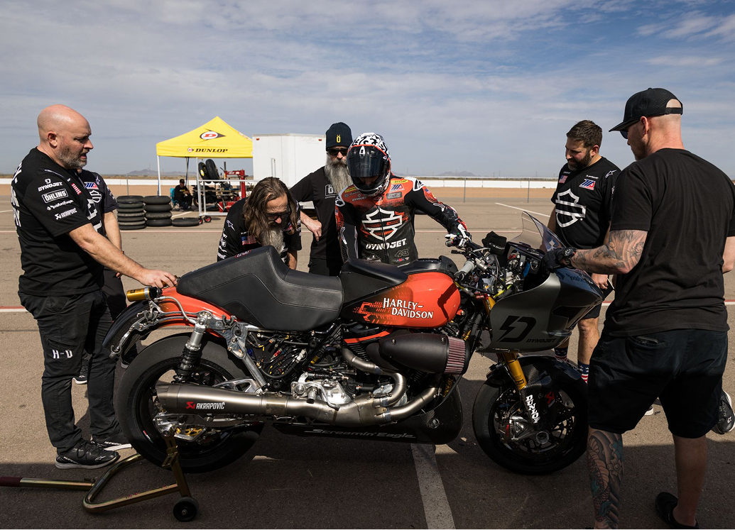 Crew members gather around a Harley-Davidson race bike as a rider prepares on a sunny track