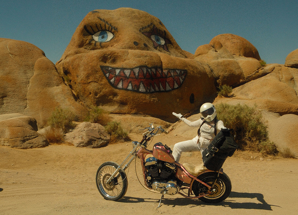 Custom chopper motorcycle and helmeted rider parked beside painted desert rock formation in a rugged landscape