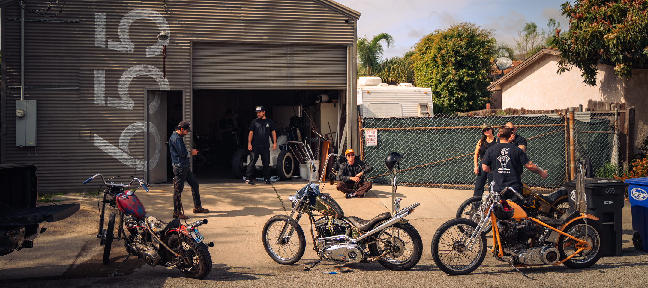 Custom motorcycles lined up outside a garage while people gather, talk, and work around the shop