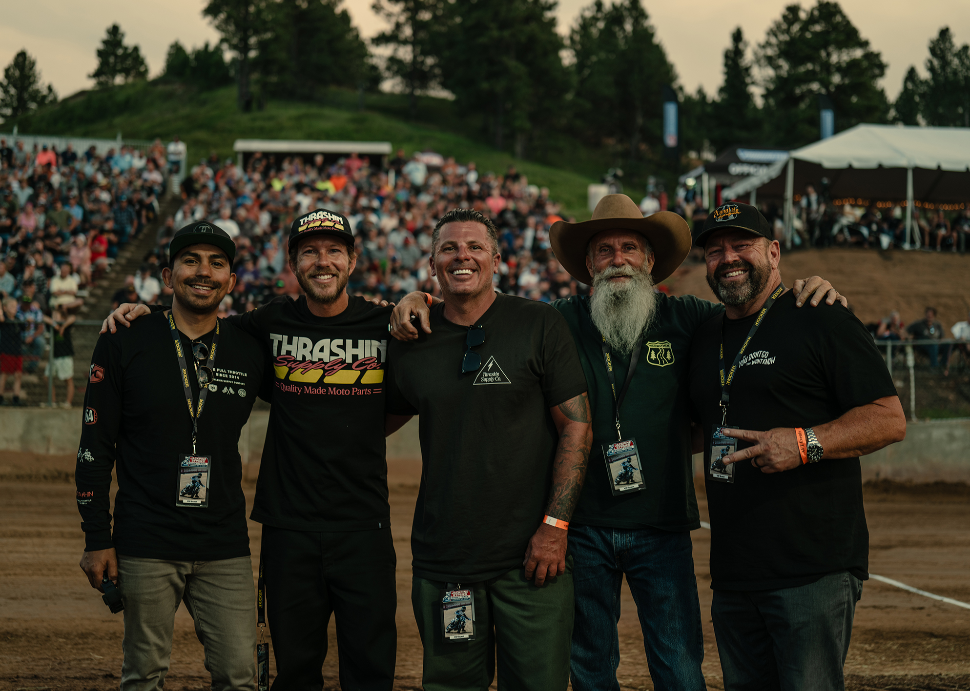 Five men pose together at a motorcycle event with a large crowd in the background.