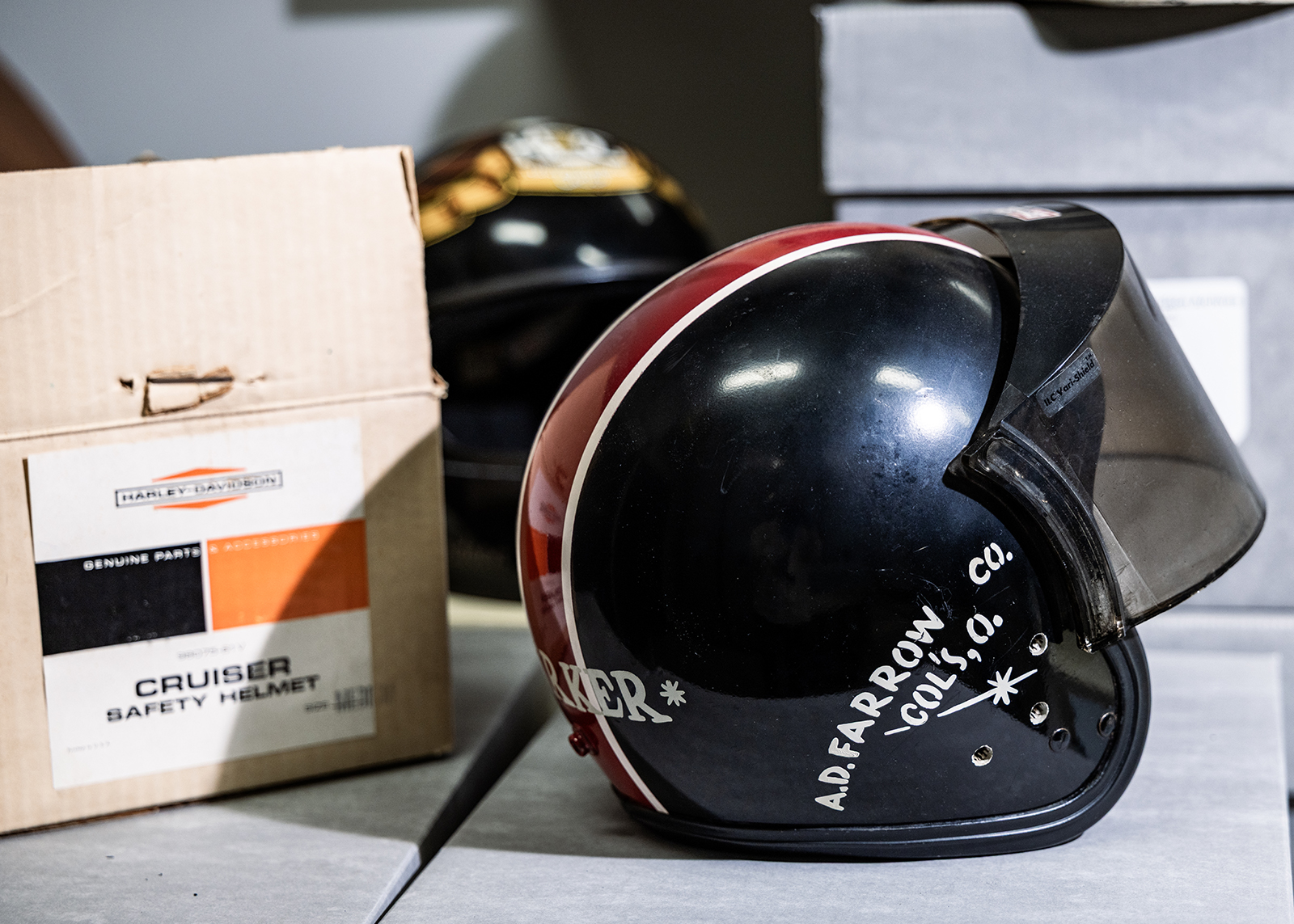 A vintage black plastic motorcycle helmet with a visor and red and white racing stripes next to Harley-Davidson Cruiser safety helmet cardboard box packaging.