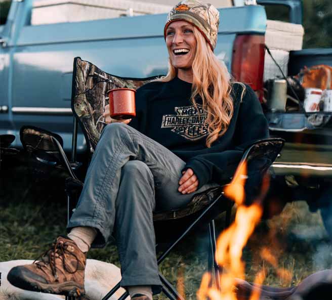 woman sitting by fire holding cup and wearing H-apparel