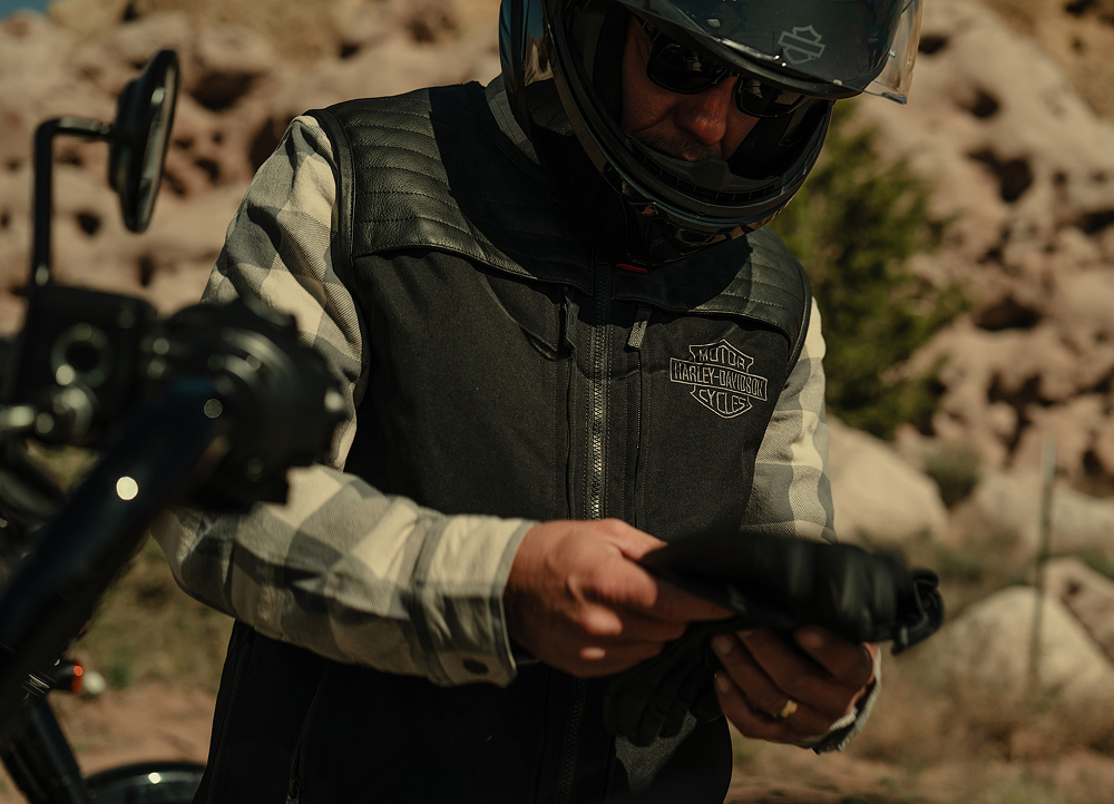 Motorcyclist wearing a Harley‑Davidson leather vest and gloves, adjusting gear beside a bike in a rocky outdoor setting