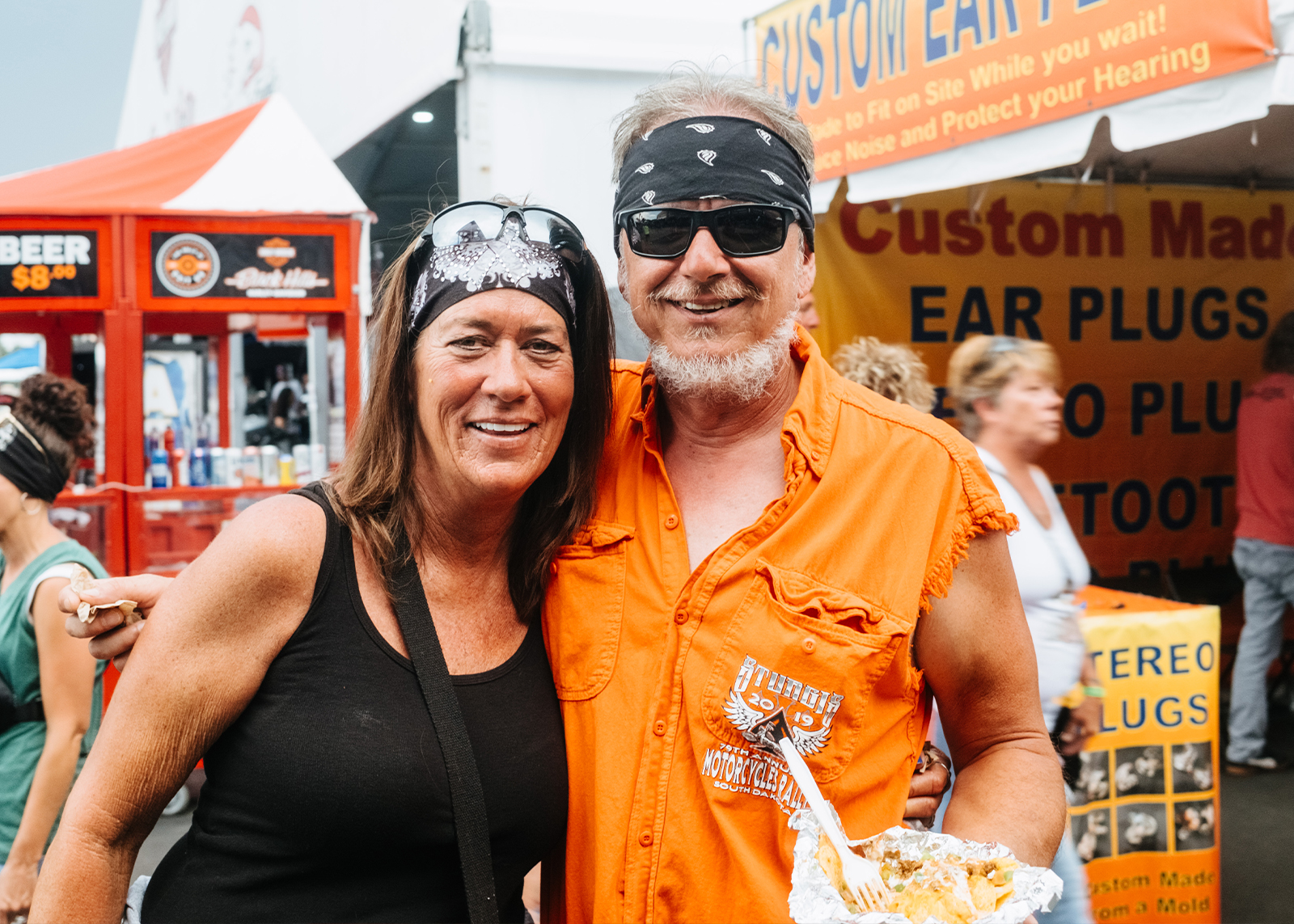 Smiling couple in biker attire pose at an outdoor rally, food in hand, with vendor tents behind them.