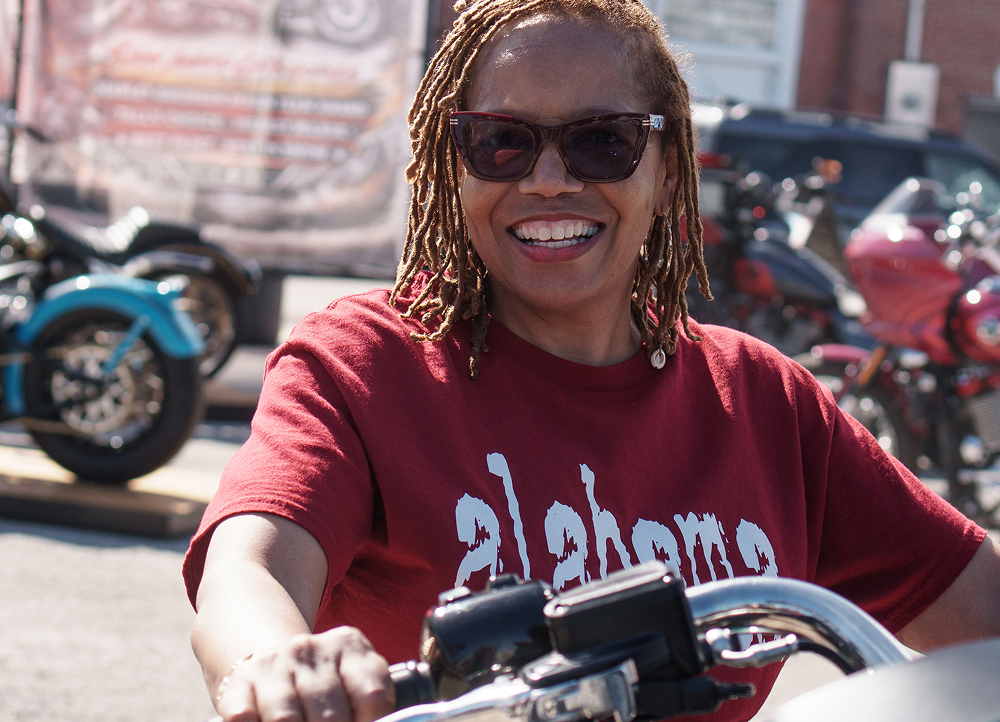Smiling person seated on a motorcycle with wide handlebars, surrounded by other bikes at an outdoor event