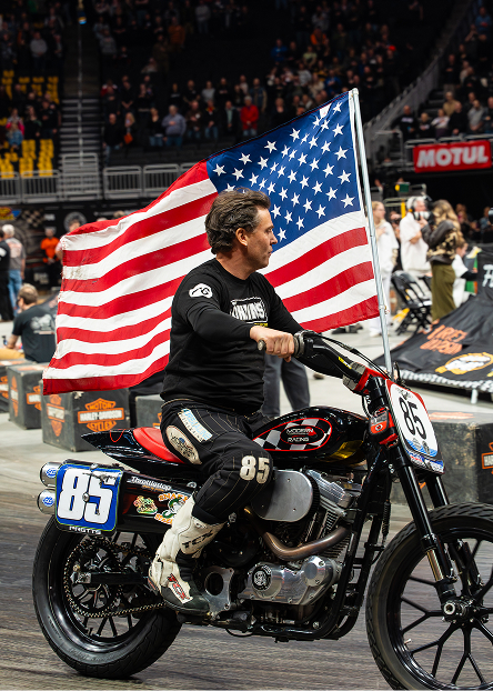 Rider on a flat-track motorcycle rides with an American flag during the Flat Out Friday event