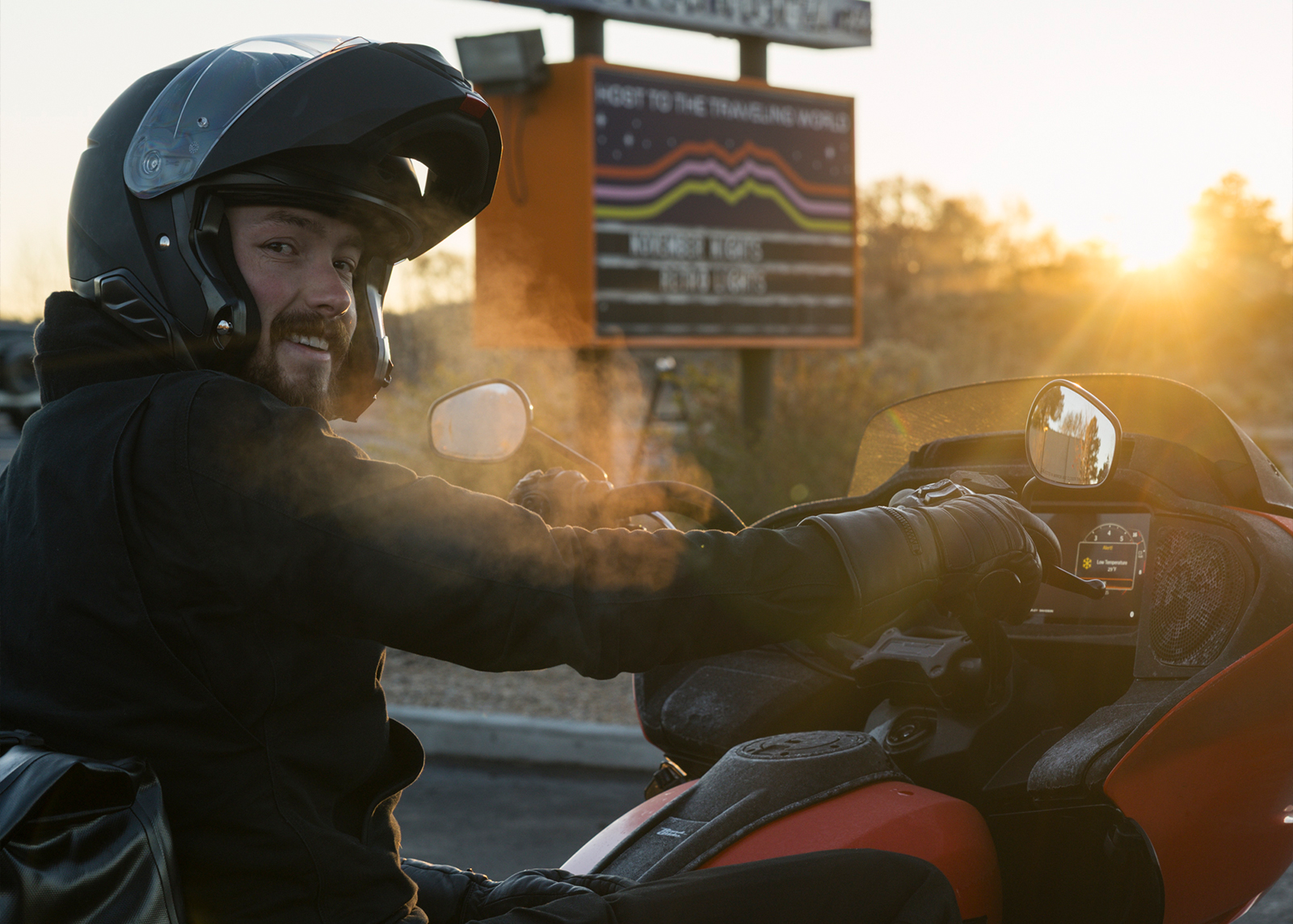A motorcyclist wearing a helmet and gloves rides at sunset, with cold fog from their breath visible in the air