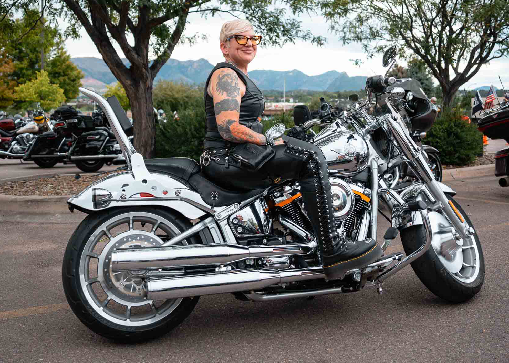 A H.O.G. member with short blonde hair, cat-eye glasses, tattoos, and full leather sits on her limited edition, all chrome Harley-Davidson Grey Ghost motorcycle.
