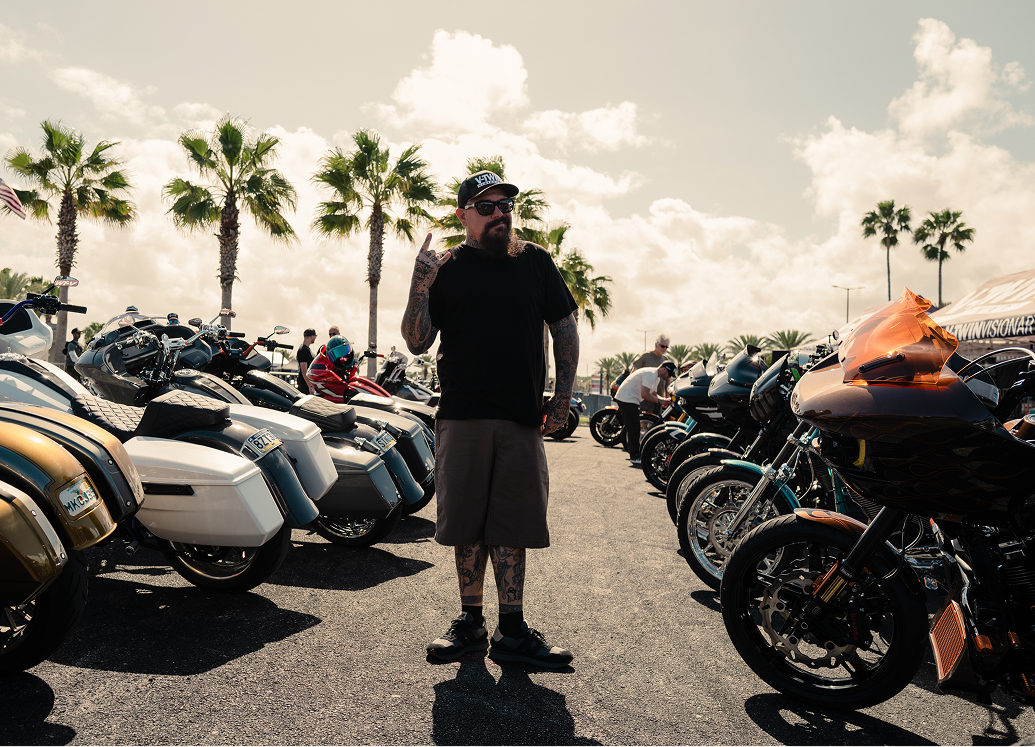 Jeff G. Holt stands between rows of parked motorcycles under palm trees at a busy Daytona rally