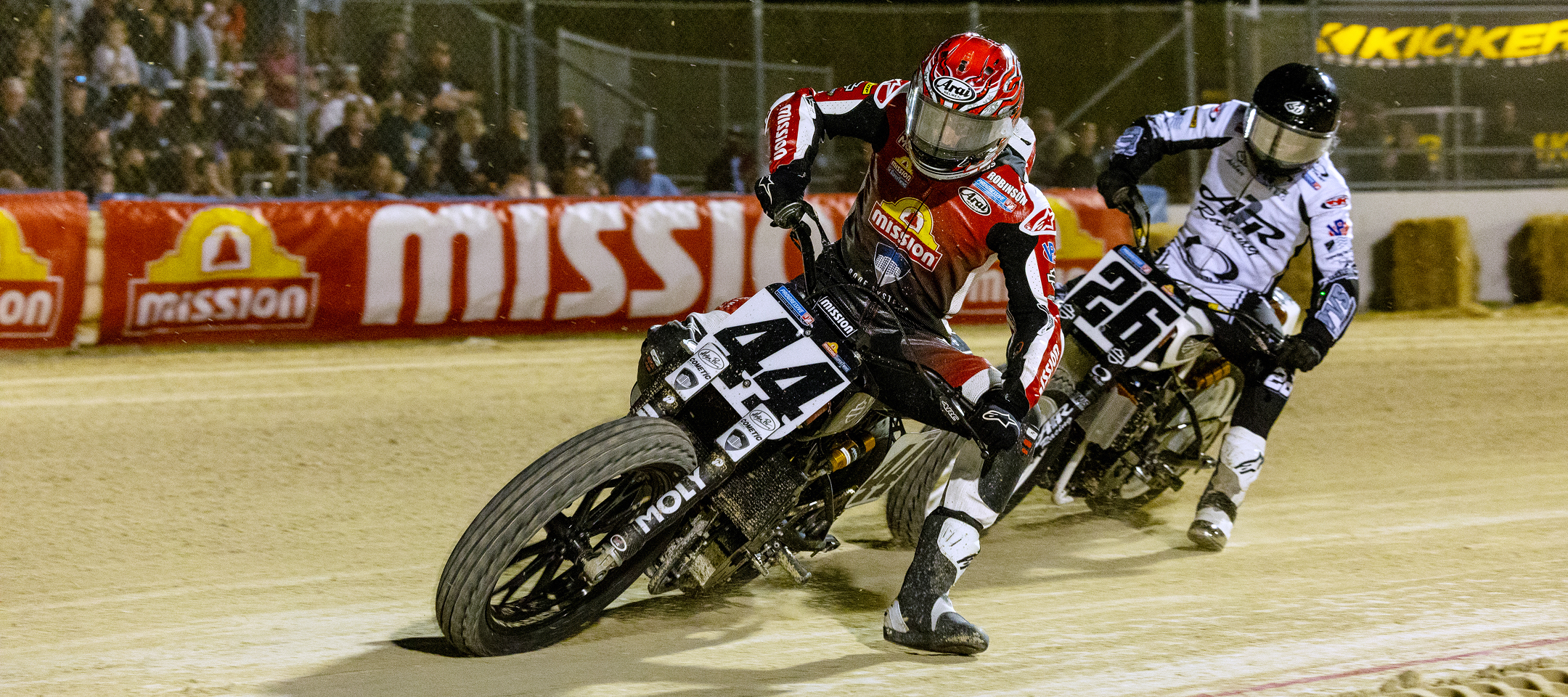 Flat track riders leaning hard into a dirt corner kicking up dust with crowd behind barrier signs glowing under lights