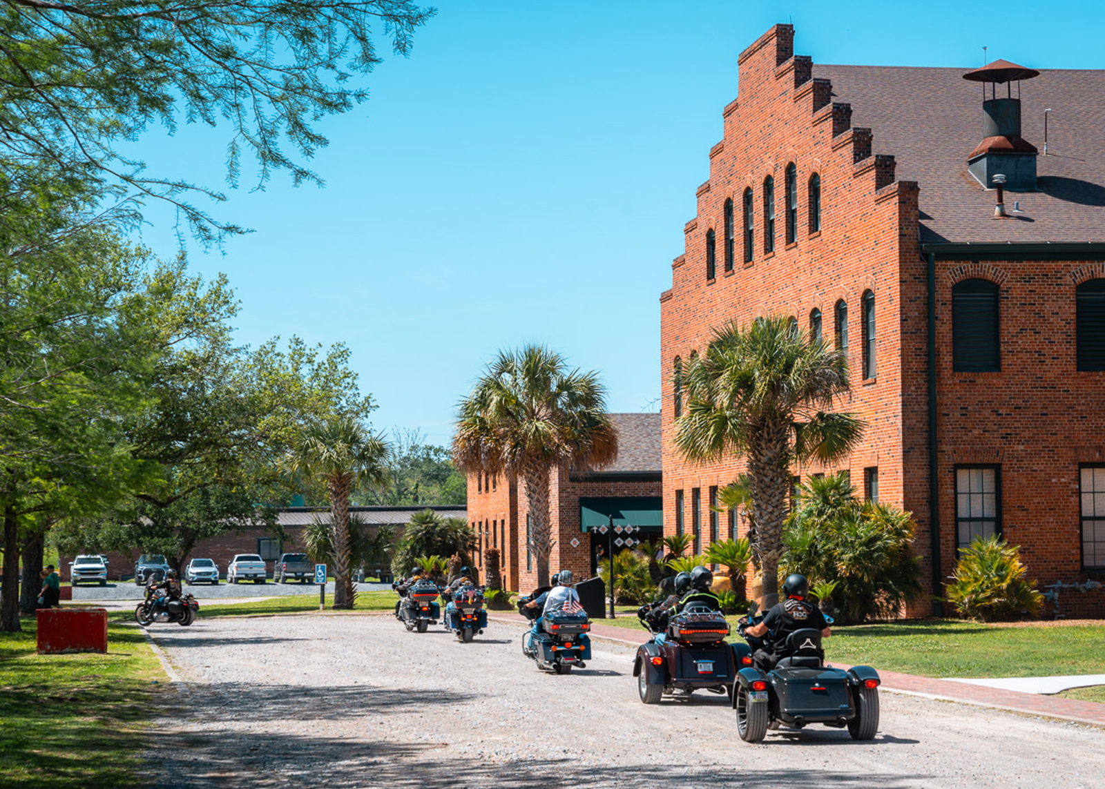 Group of motorcycles riding past a historic red brick building with palm trees on a sunny day