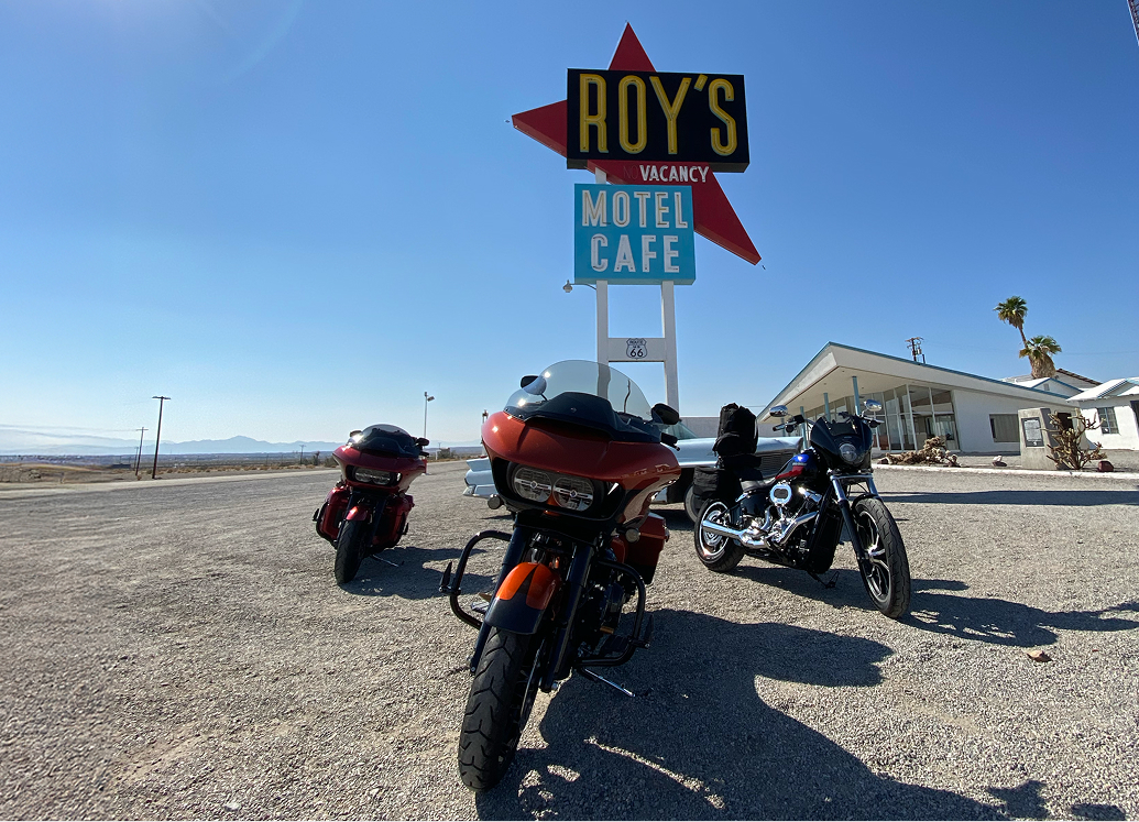 Three motorcycles parked in front of a retro Roy’s Motel Café sign under a clear sky in a desert landscape