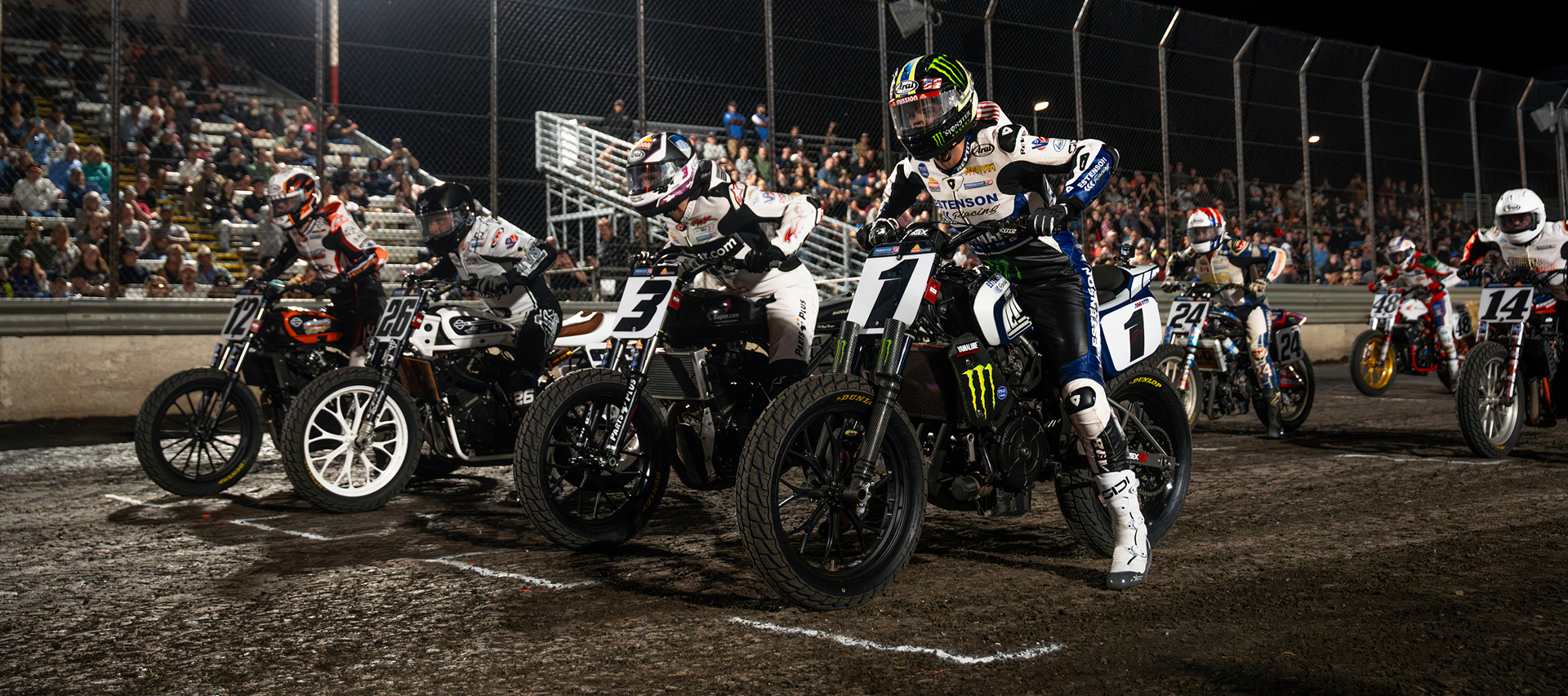 Flat track motorcycle racers launch from the starting line during a nighttime race as spectators fill the grandstands behind the dirt track