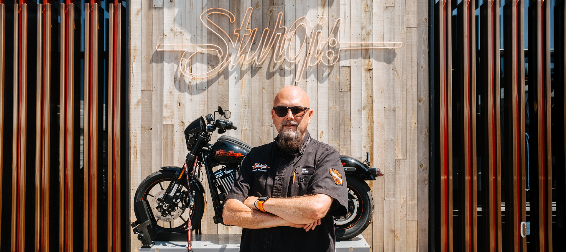 Sturgis Harley-Davidson’s General Manager Tim Sutherland wearing sunglasses and Harley-Davidson shirt poses in front of motorcycle and neon Sturgis sign.