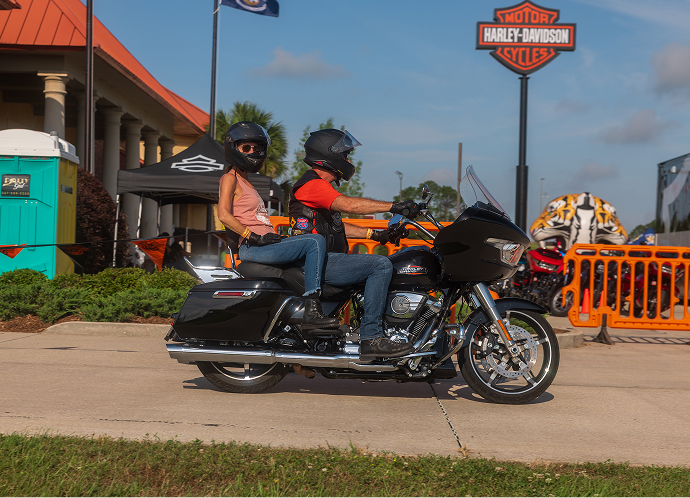 Two riders pass on a touring motorcycle outside a Harley-Davidson dealership on a sunny day