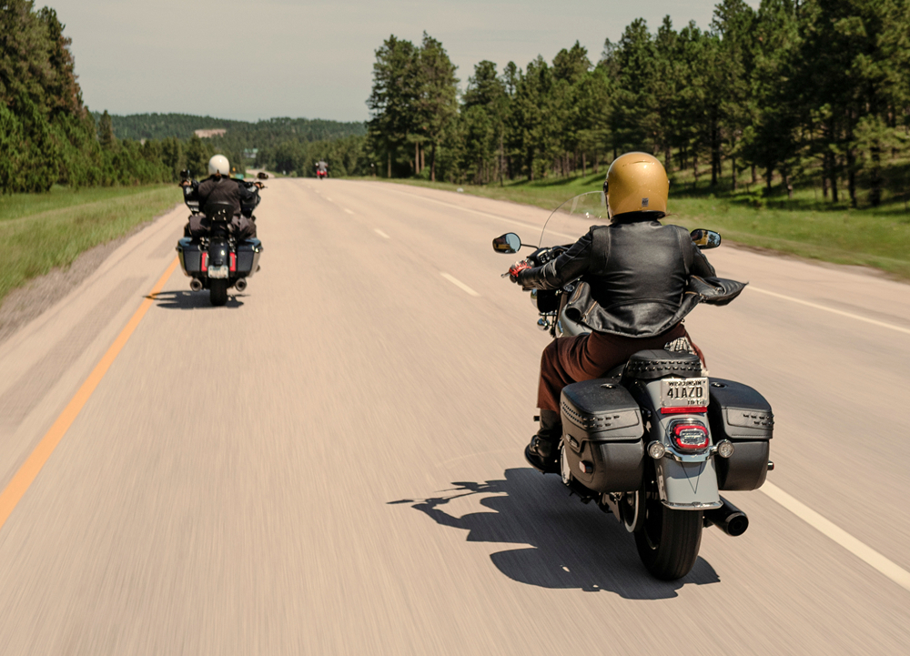 Two H-D riders cruising on open highway with rolling hills and pine trees under clear sky on scenic ride.
