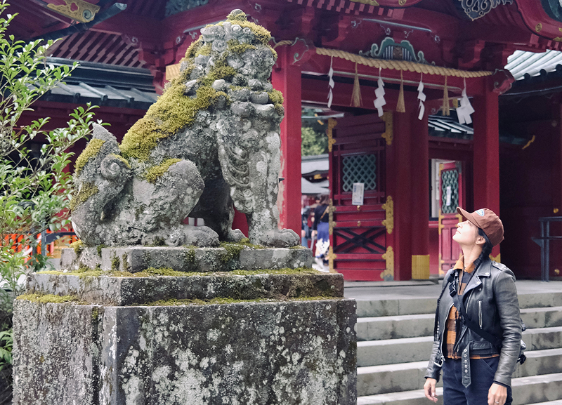 Adri Law standing beside a moss-covered stone guardian statue in front of a red temple structure with steps and greenery nearby