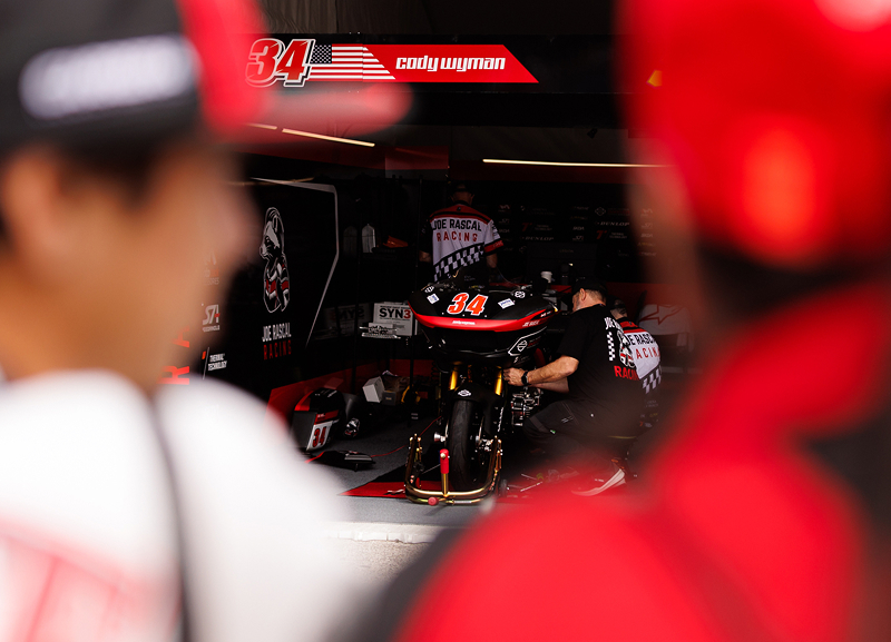Spectators look into a race team garage as crew members prepare a motorcycle before competition