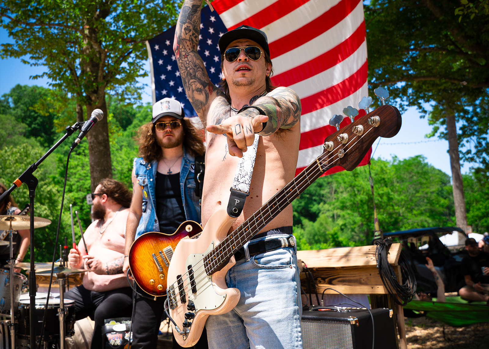 A shirtless man with his guitar points towards the camera with his band on stage behind him.