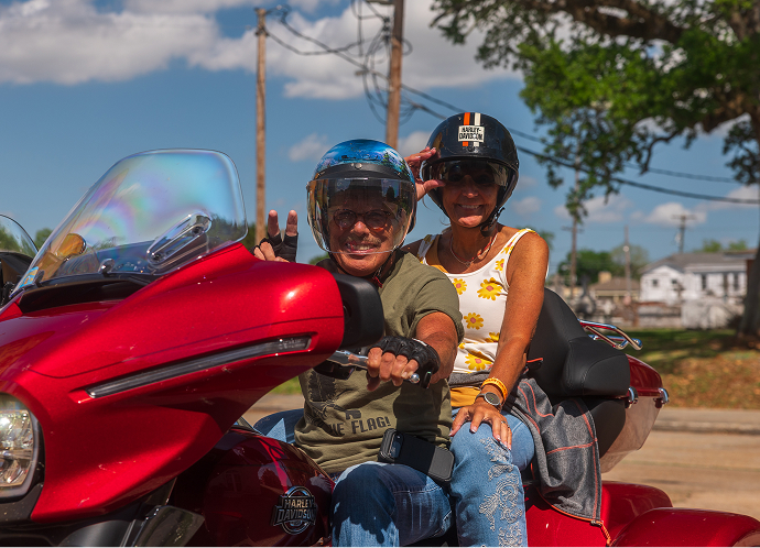 Two riders sit together on a red touring motorcycle, parked and waving at the camera during a group ride event