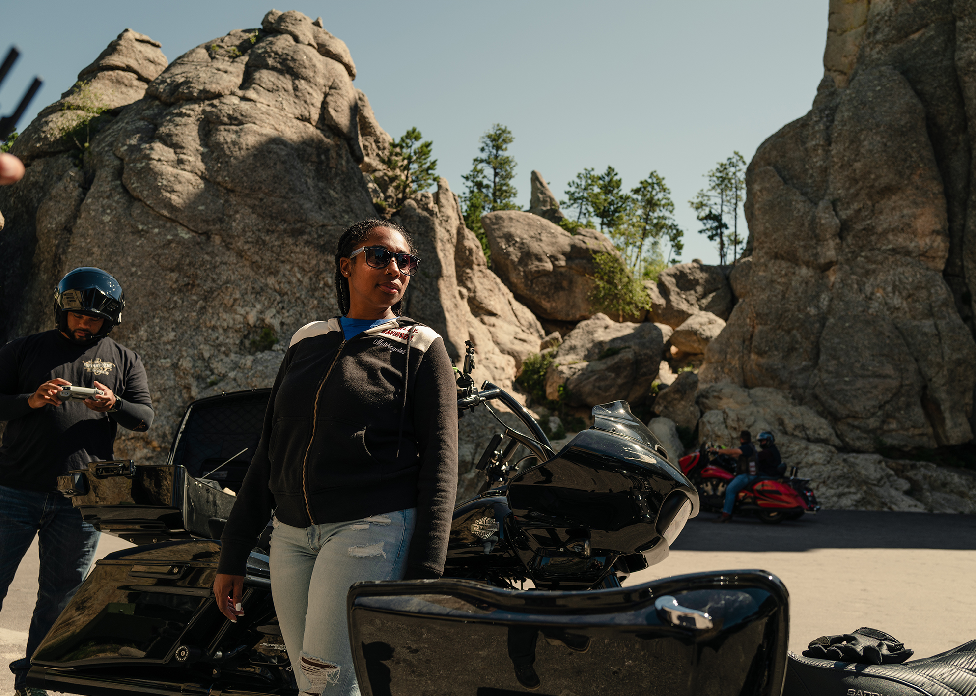 Woman in sunglasses stands beside a black Harley-Davidson motorcycle amid large rock formations.