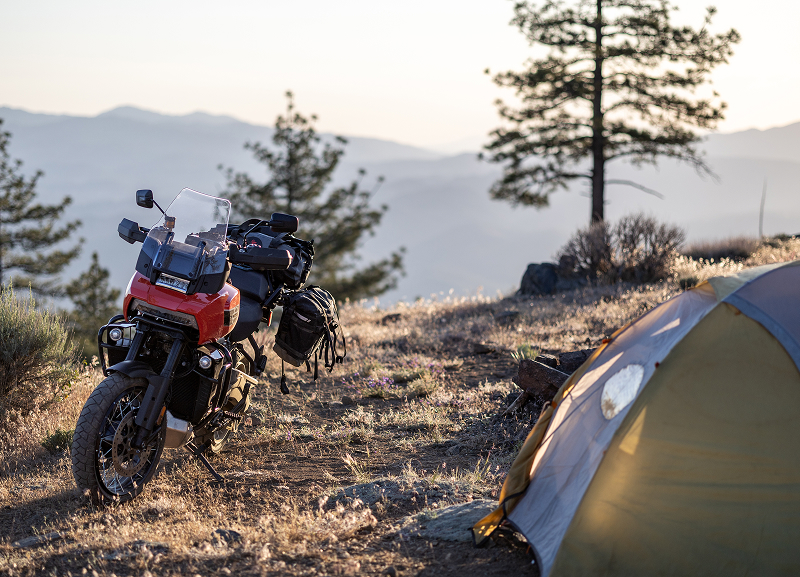 Packed Red H-D Pan America motorcycle parked beside camping tent on hillside with trees and distant mountains