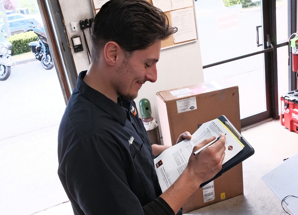 Precision H-D employee reviewing paperwork on clipboard with shipping box and glass door in background