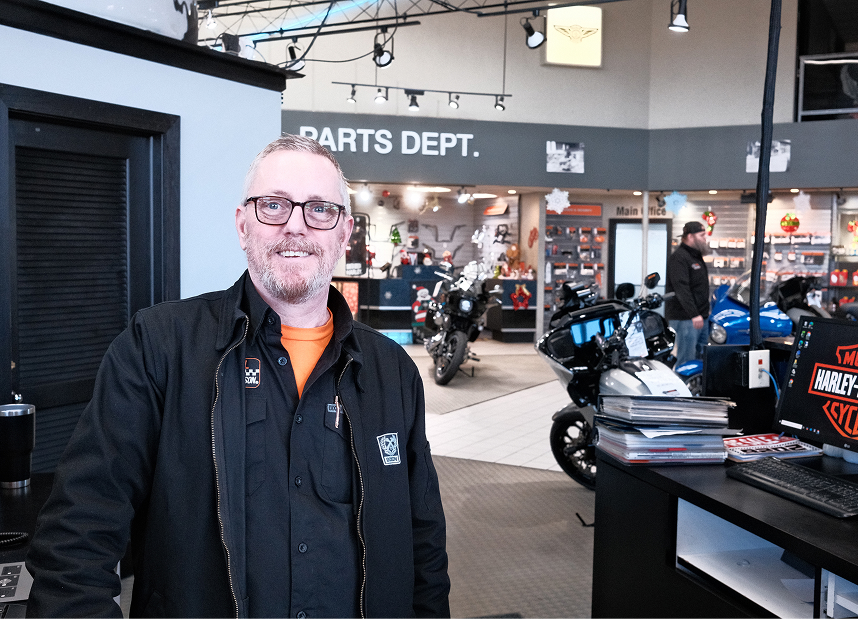 An employee with glasses and a mechanics jacket poses for a photo in the showroom of Kegel Harley-Davidson