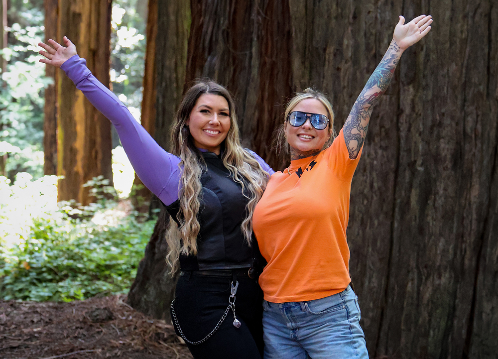 Two travelers pose beneath towering redwoods, dwarfed by the immense trees in the forest