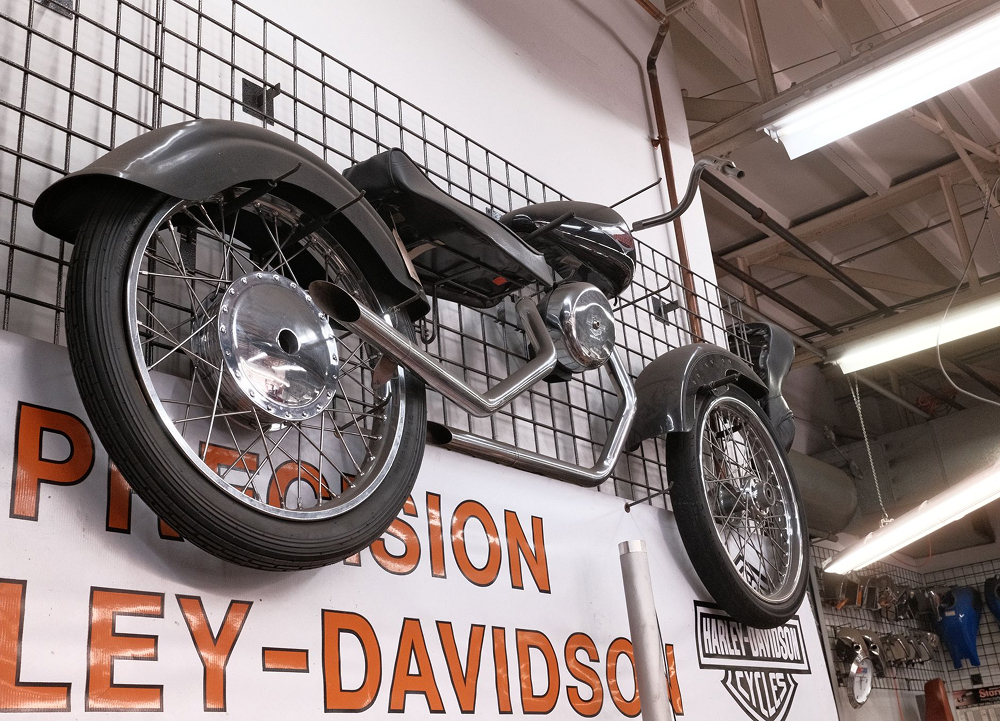 Low angle view of deconstructed motorcycle mounted above Precision H-D sign with workshop lighting and metal grid backdrop