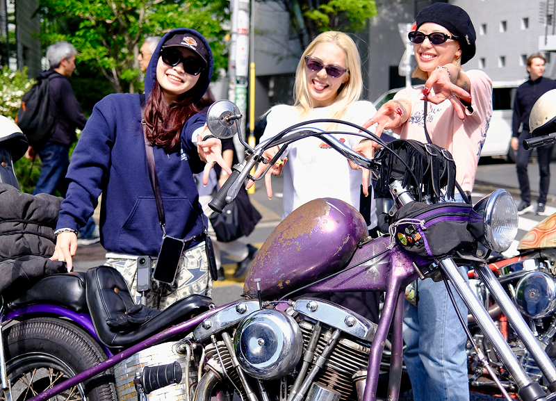Three riders gathered around a purple custom motorcycle parked on a city street, pointing toward camera with buildings behind