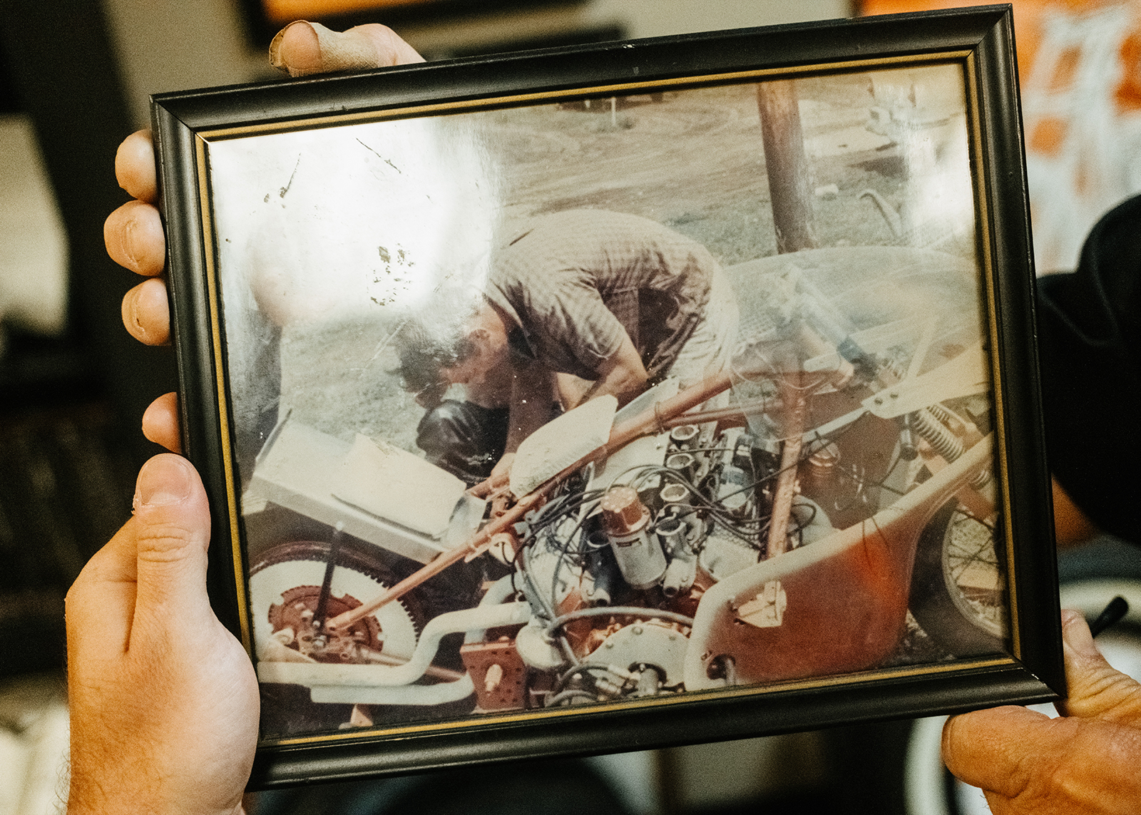 A framed, vintage photo of a man working on a motorcycle being held up for the camera.