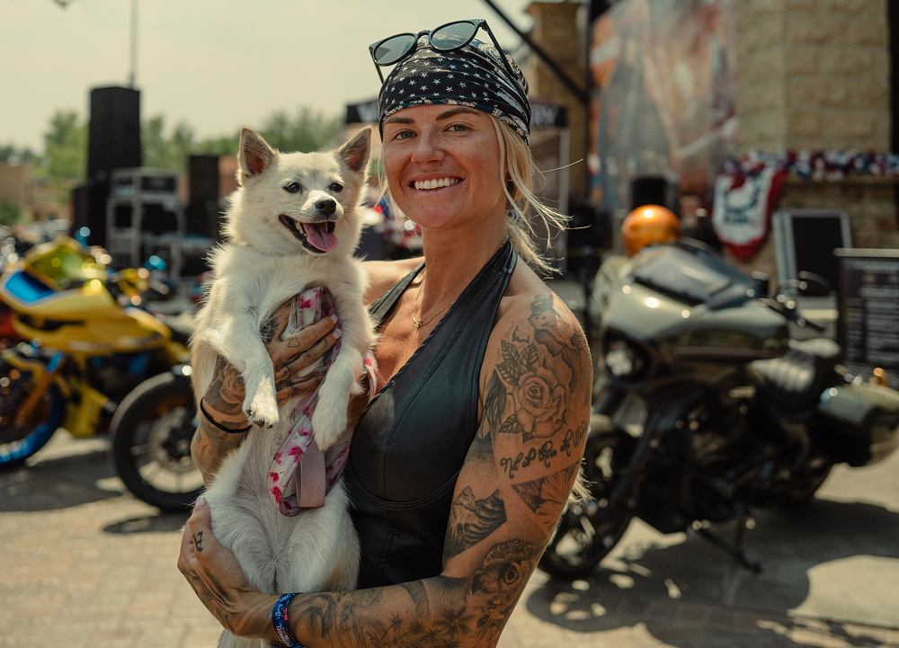 Person holding a small white dog among custom motorcycles and crowds at a busy Sturgis rally event