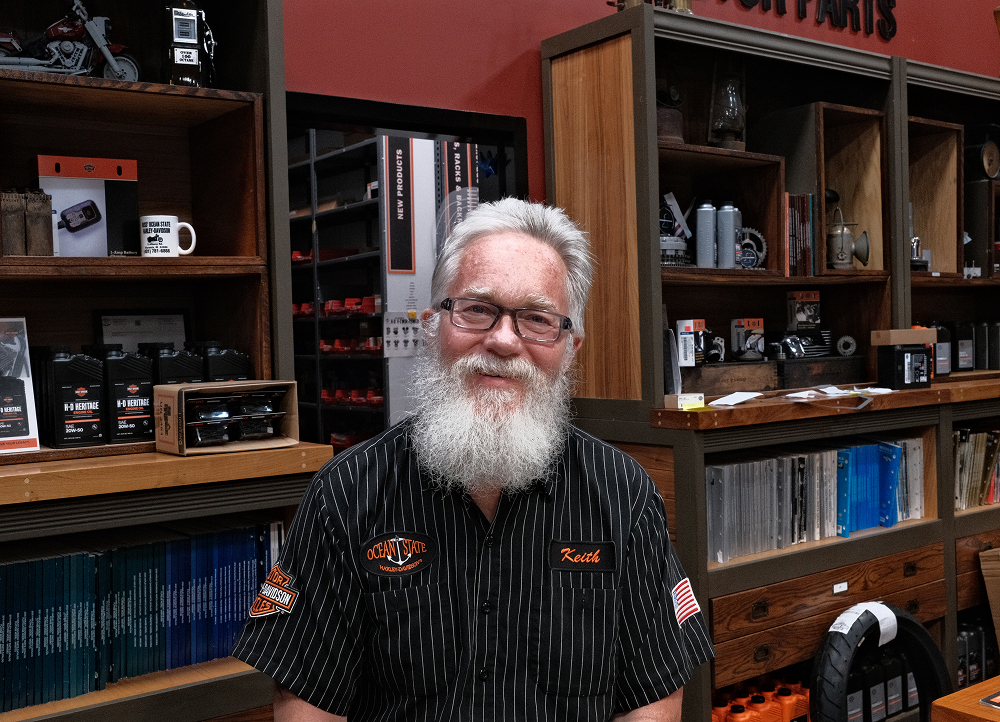 Staff member with grey hair and beard surrounded by shelves of tools, manuals, and Harley-Davidson products
