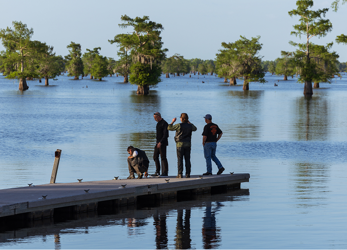 Group of riders stand and crouch on a dock extending into a flooded cypress swamp with trees rising from the water
