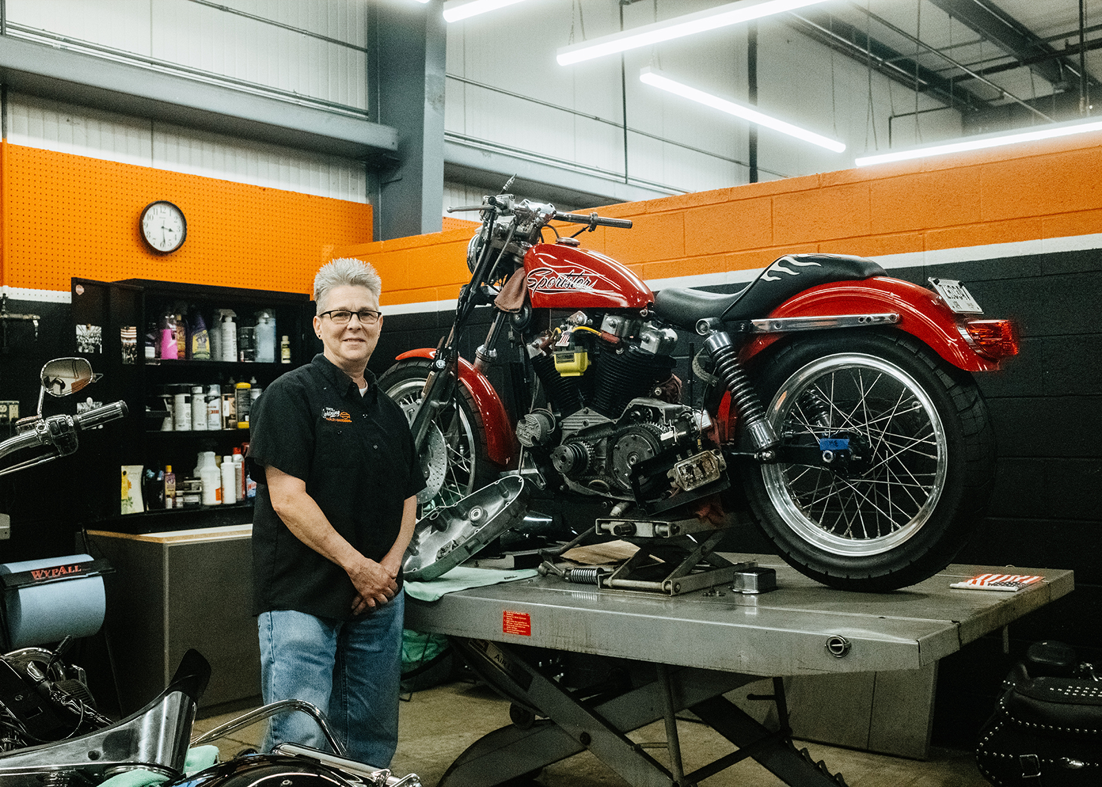 A service technician poses next to a Sportster on a lift that is being worked on.