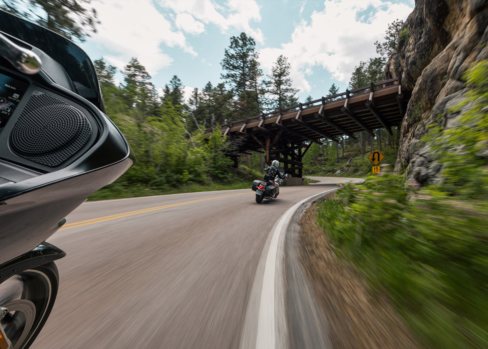 Motorcycles leaning into a curve on a scenic forest road beneath a wooden bridge structure