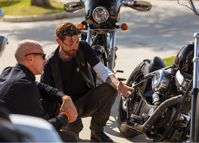 Two riders crouch beside a parked motorcycle, inspecting details and talking in a parking lot