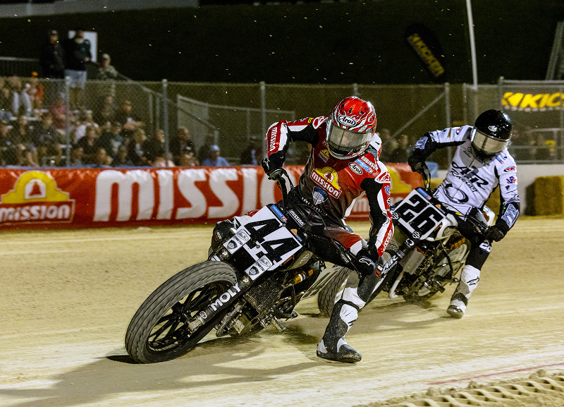 Flat track riders leaning hard into a dirt corner kicking up dust with crowd behind barrier signs glowing under lights