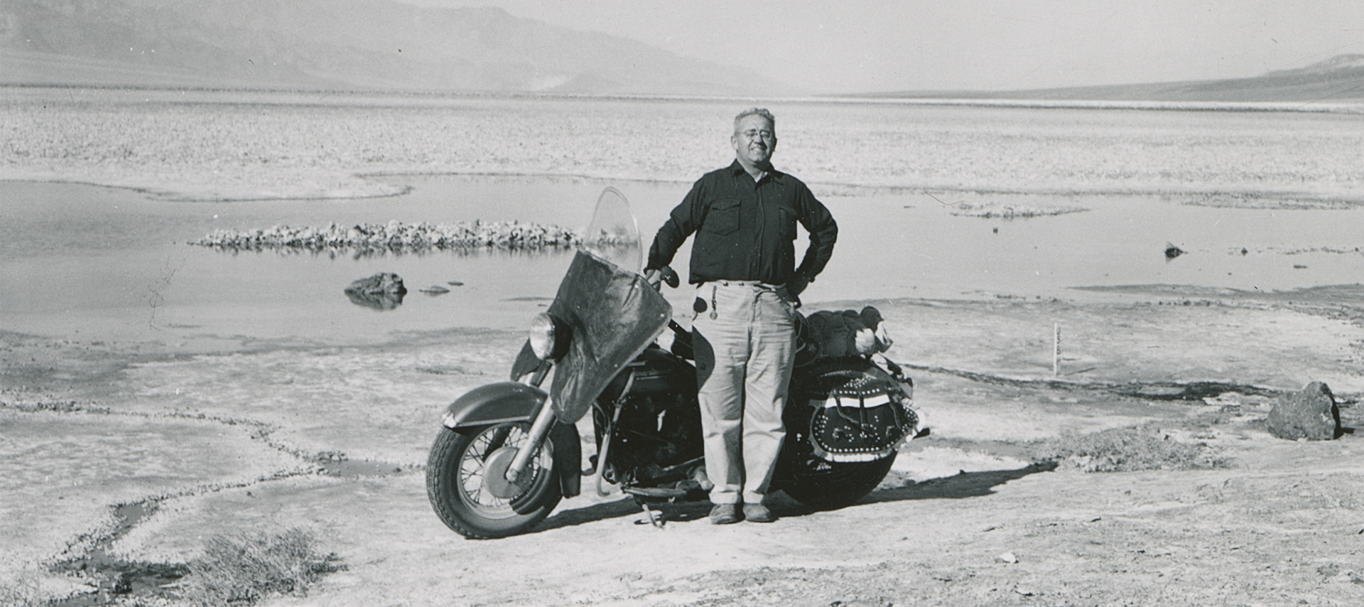 Black and white historical photo of Eldred Harrington standing by his 1951 Harley-Davidson Hydra Glide motorcycle in the desert.