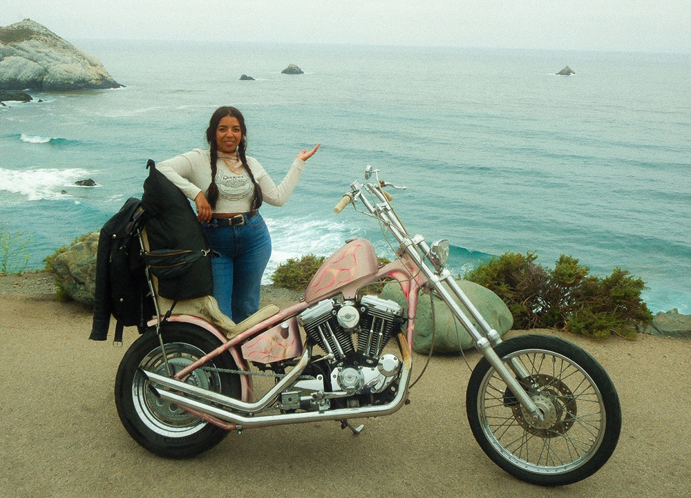 Custom chopper-style motorcycle parked at a coastal overlook with ocean waves, rocky shoreline, and rider beside bike