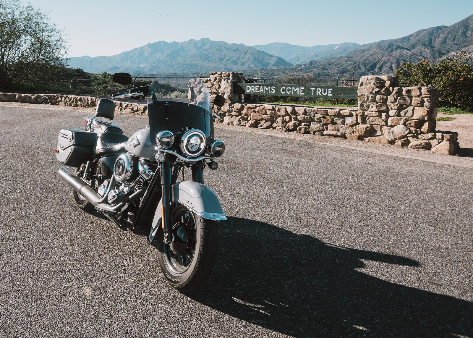 A Harley-Davidson Heritage Classic motorcycle parked at a scenic overlook with stone wall and wooden sign that says “Dreams come true” in the background